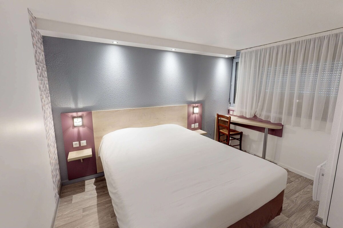 Une chambre d’hôtel avec un grand lit, un bureau avec une chaise, une fenêtre avec des rideaux légers et un luminaire fixé au mur.