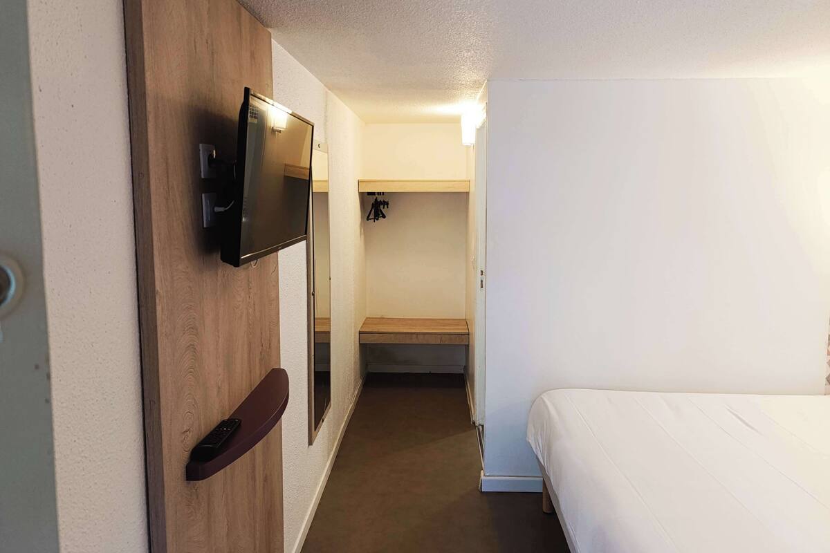 Et hotelværelse med en trævæg, et ophængt tv, en seng og en lille træhylde med en fjernbetjening.