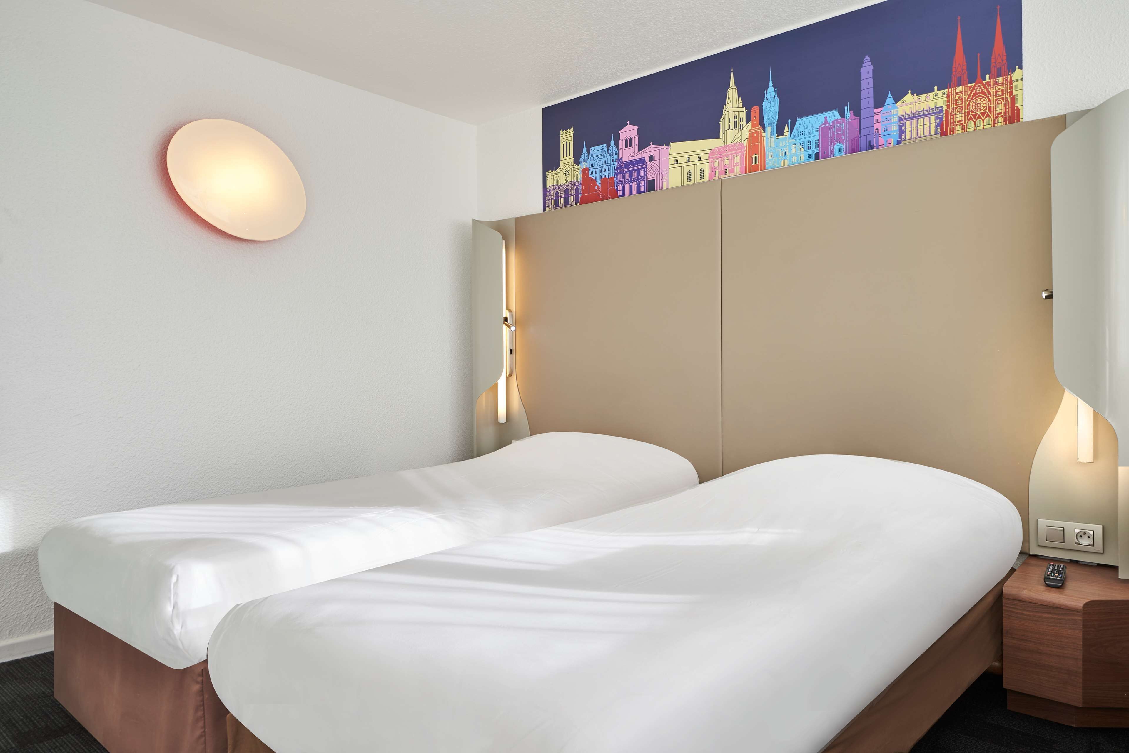 Habitació d'hotel amb dos llits, una obra d'art amb un paisatge urbà a la paret, una làmpada rodona i una tauleta de nit amb una làmpada.