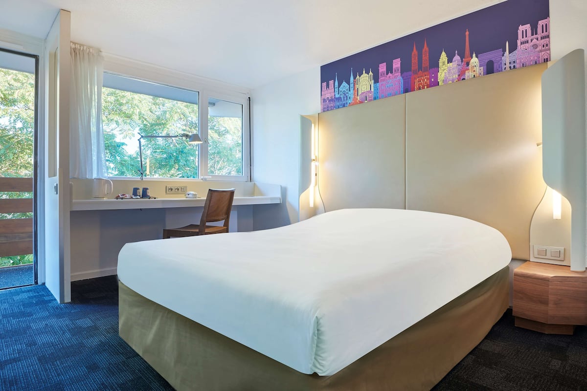Ein Hotelzimmer mit einem großen Bett, einem Schreibtisch mit Stuhl, einem Fenster und einem Wandgemälde mit Stadtansicht.