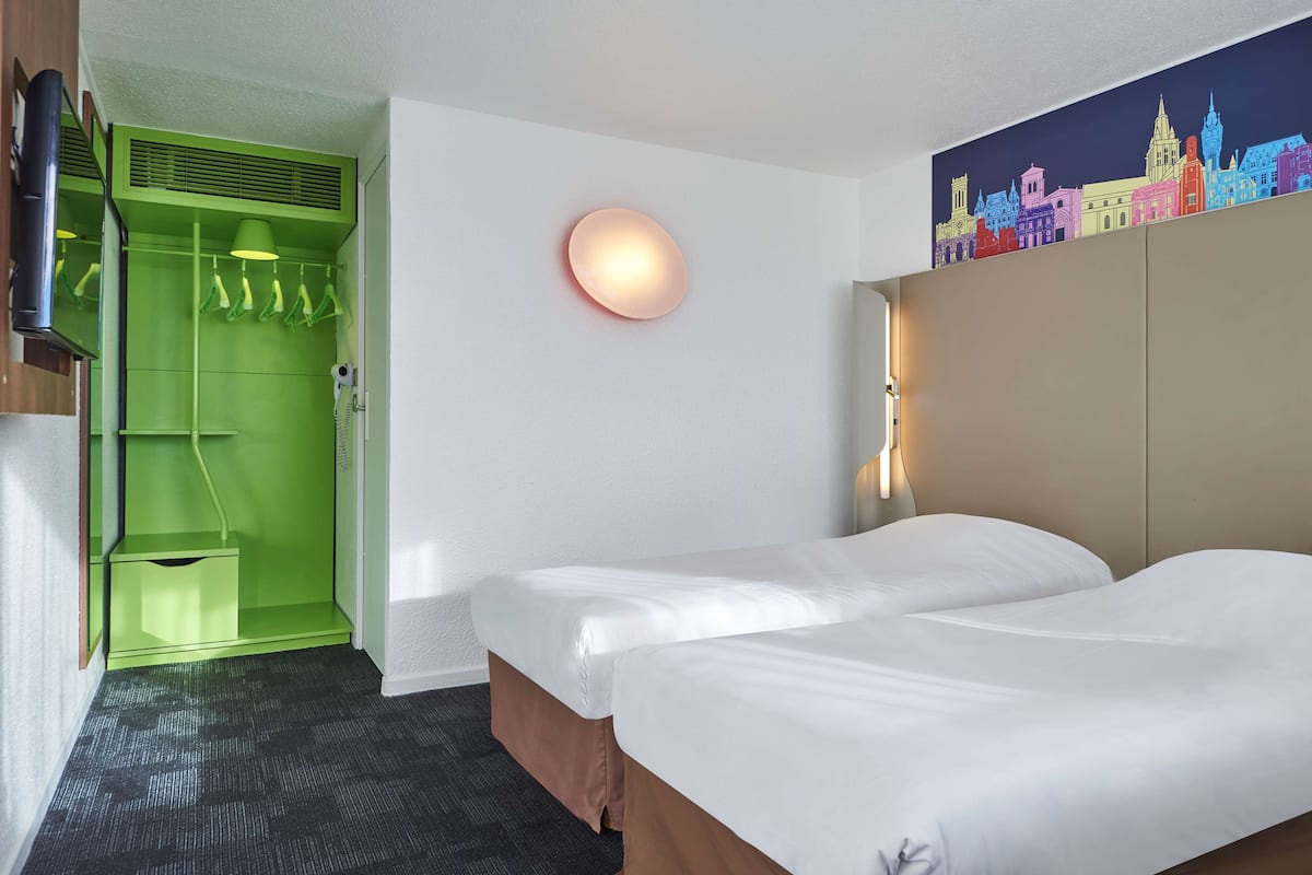 Ein Hotelzimmer mit zwei Betten, einem grünen Schrank, einem Stadtbild-Wandgemälde und einer runden Wandleuchte.