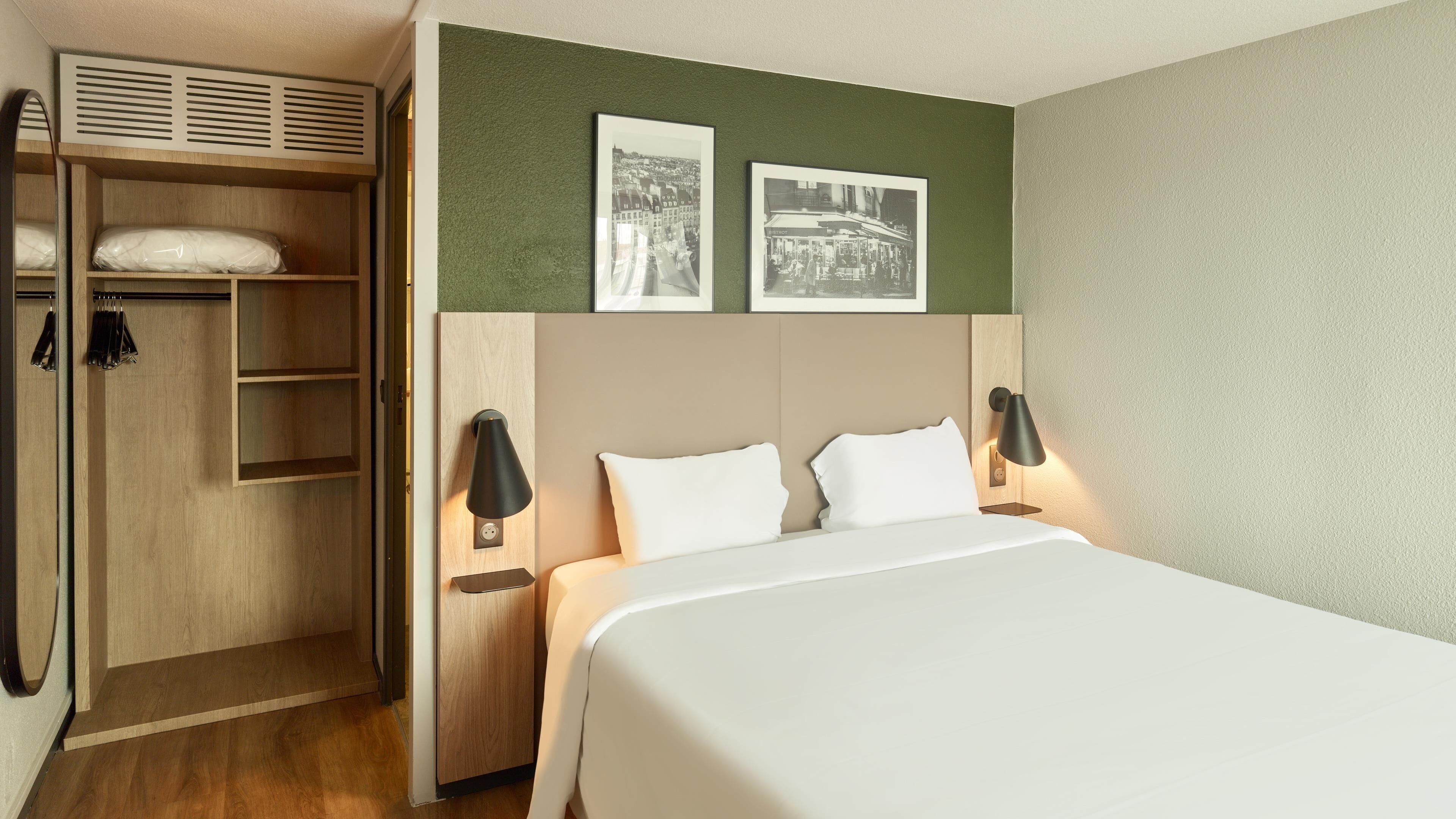 Quarto de hotel com cama, duas luminárias de cabeceira, um guarda-roupa e duas molduras na parede.