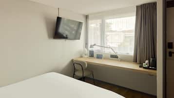 A hotel room with a bed, a desk with a chair, a TV mounted on the wall, a window with curtains, and a thermostat on the window sill.