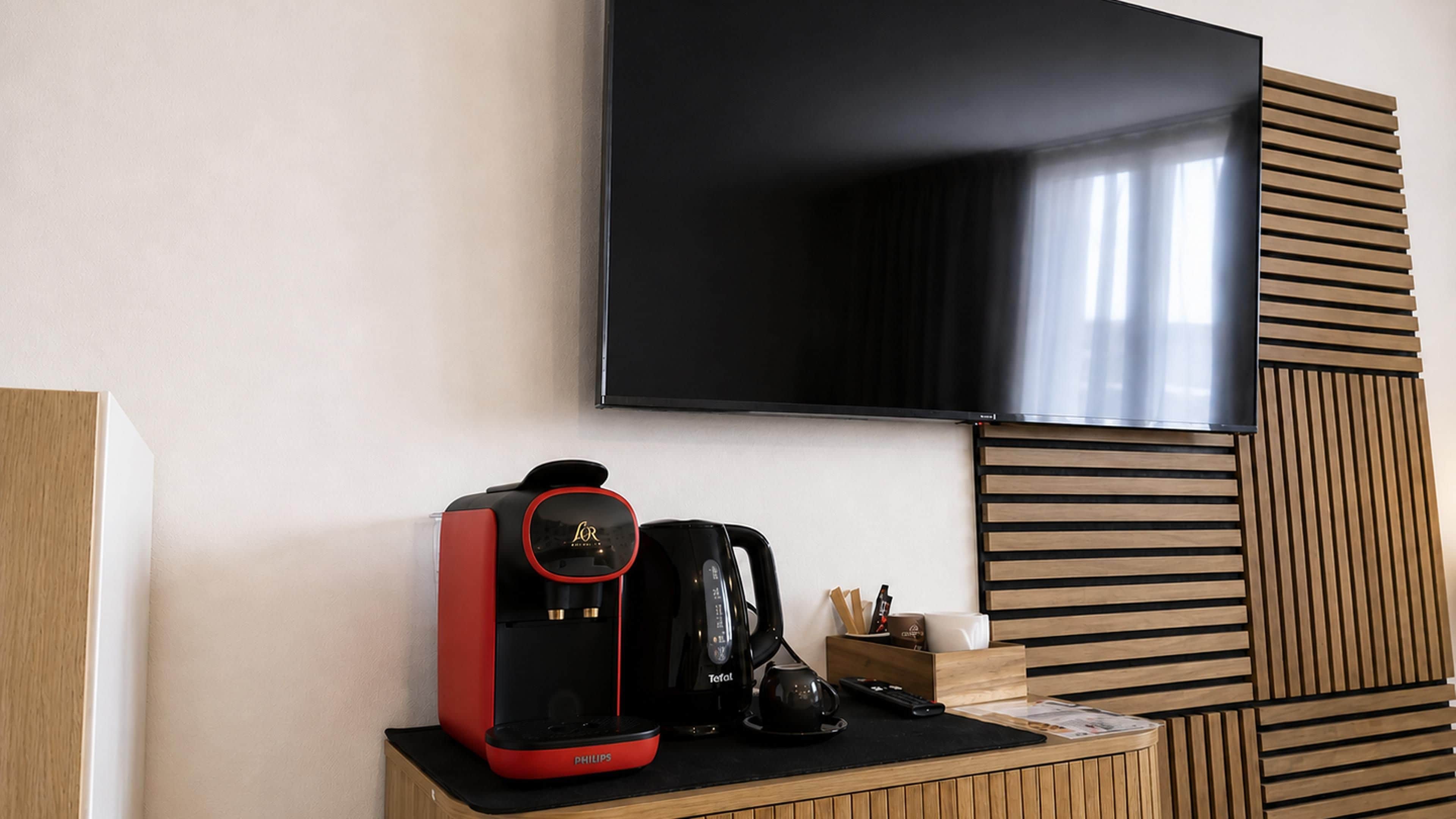 Ein modernes Hotelzimmer mit einem an der Wand befestigten Flachbildfernseher, einer roten und schwarzen Kaffeemaschine auf einer Holztruhe und einem Holzschrank mit Lamellen.