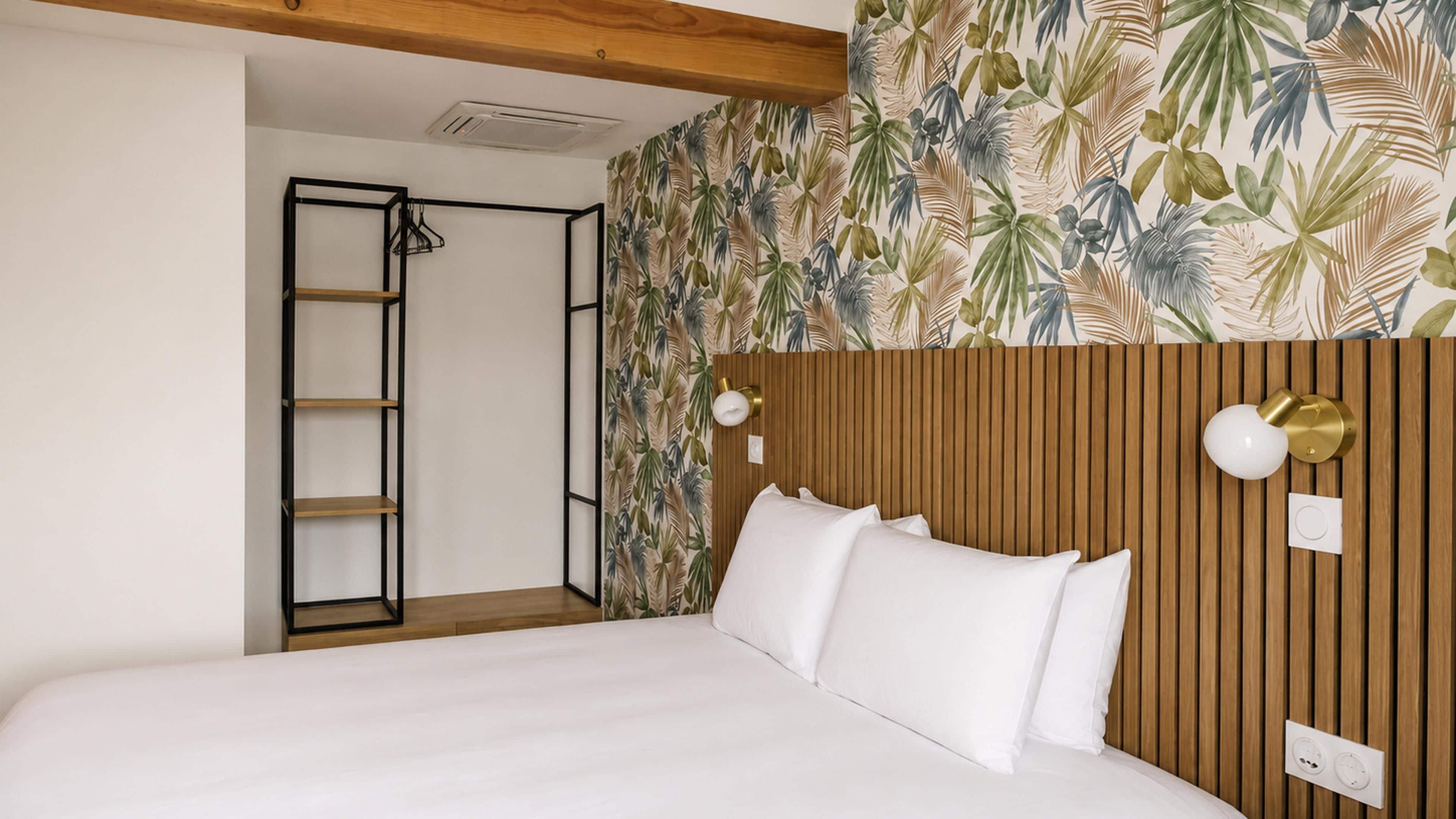 Une chambre d’hôtel avec un lit, une tête de lit en bois, un mur orné d’un motif de feuilles tropicales, une échelle en métal noir et un luminaire fixé au mur.