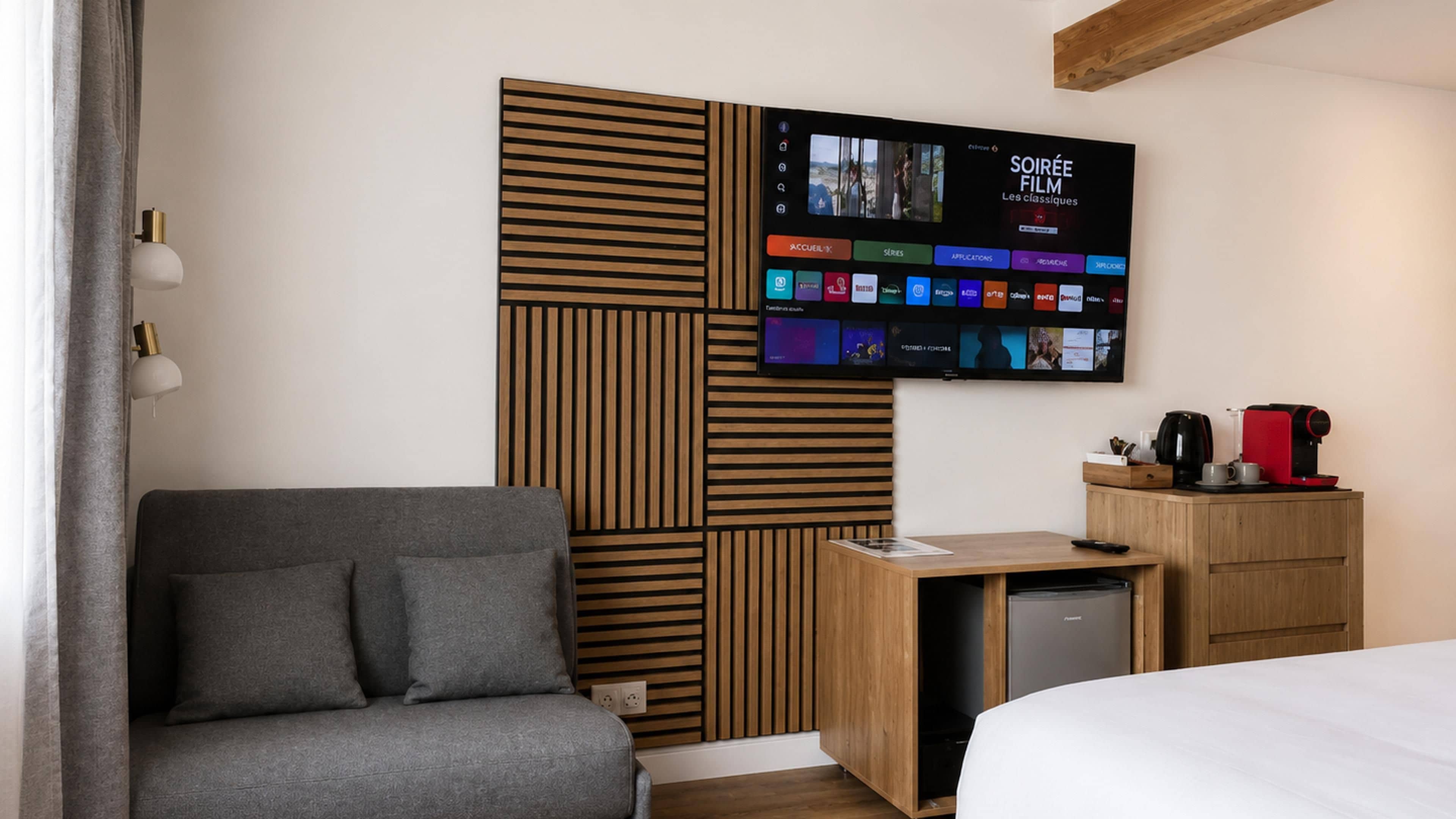 Habitación de hotel con una televisión de pantalla plana montada en la pared mostrando un canal de cine, un sofá gris, un mueble de madera con una cafetera y una cama con ropa blanca.