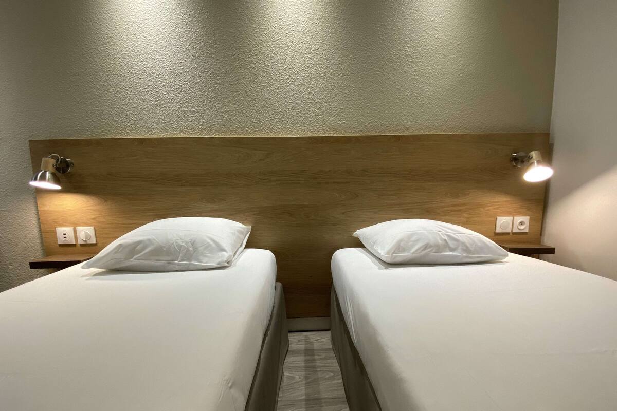Habitación de hotel con dos camas individuales, cada una con una almohada blanca y un cabecero de madera. Hay lámparas de noche a ambos lados de las camas.