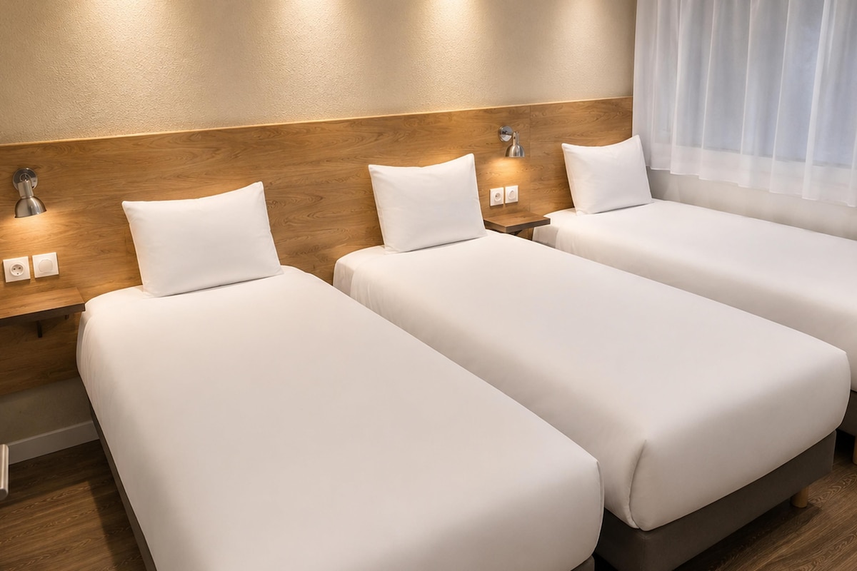A hotel room with three single beds, wooden headboards, bedside lamps, and a window with curtains.