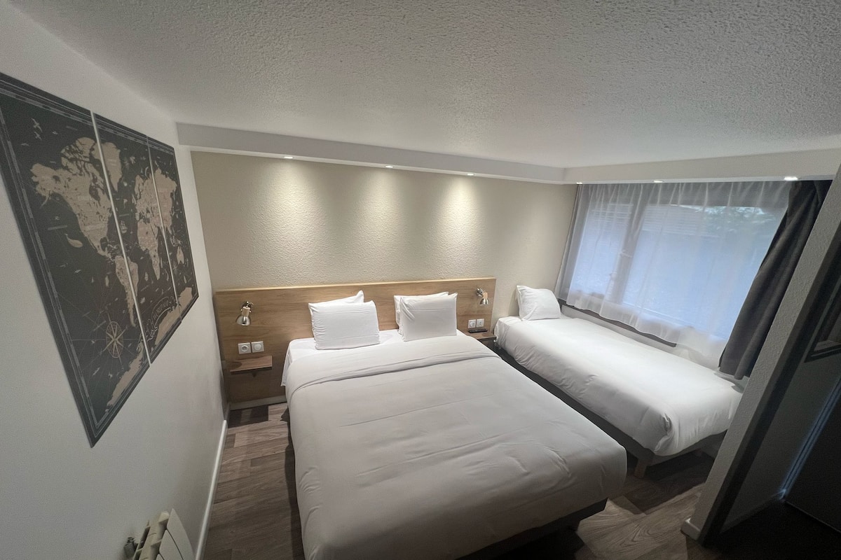 Habitación de hotel con dos camas, cabecera de madera, mesita de noche, lámpara de pared y un gran ventanal con cortinas traslúcidas.