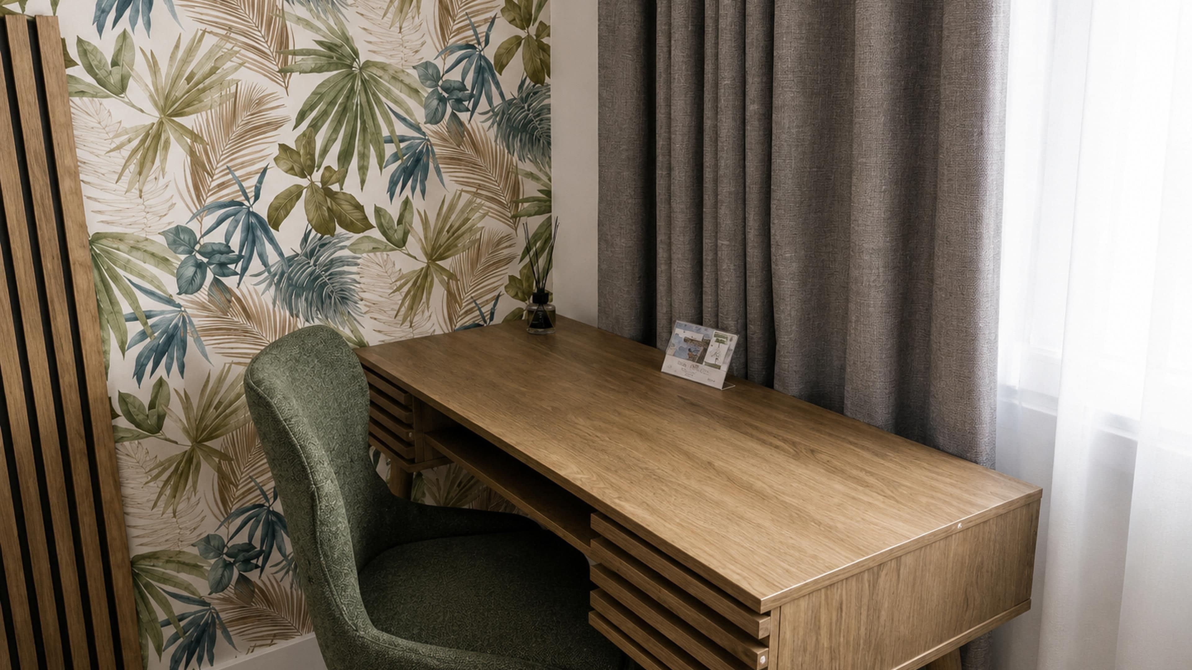 A hotel room with a wooden desk, a green chair, a tropical leaf wallpaper, and grey curtains.