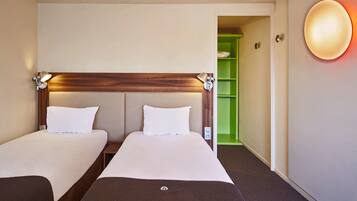 A hotel room with two beds, a wooden headboard, a green shelving unit, and a wall-mounted light fixture.