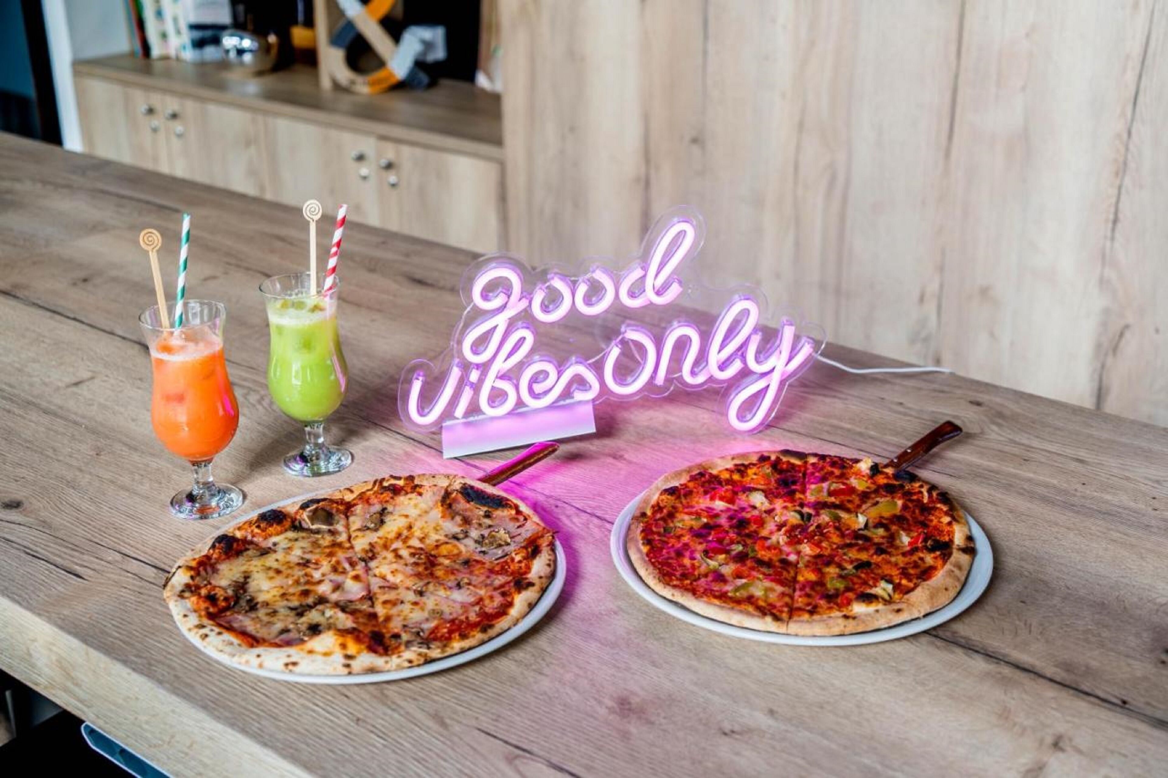Zwei Pizzen auf einem Holztisch, mit einem Neon-Schild, das 'Good Vibes Only' zeigt, und zwei Getränke mit Strohhalmen.