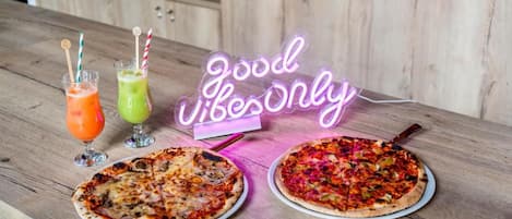 Dos pizzas sobre una mesa de madera con un letrero de neón que dice 'Good Vibes Only' y dos bebidas con pajitas.