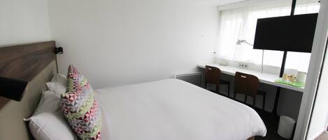 A hotel room with a bed, a desk, a chair, a lamp, and a window with curtains.