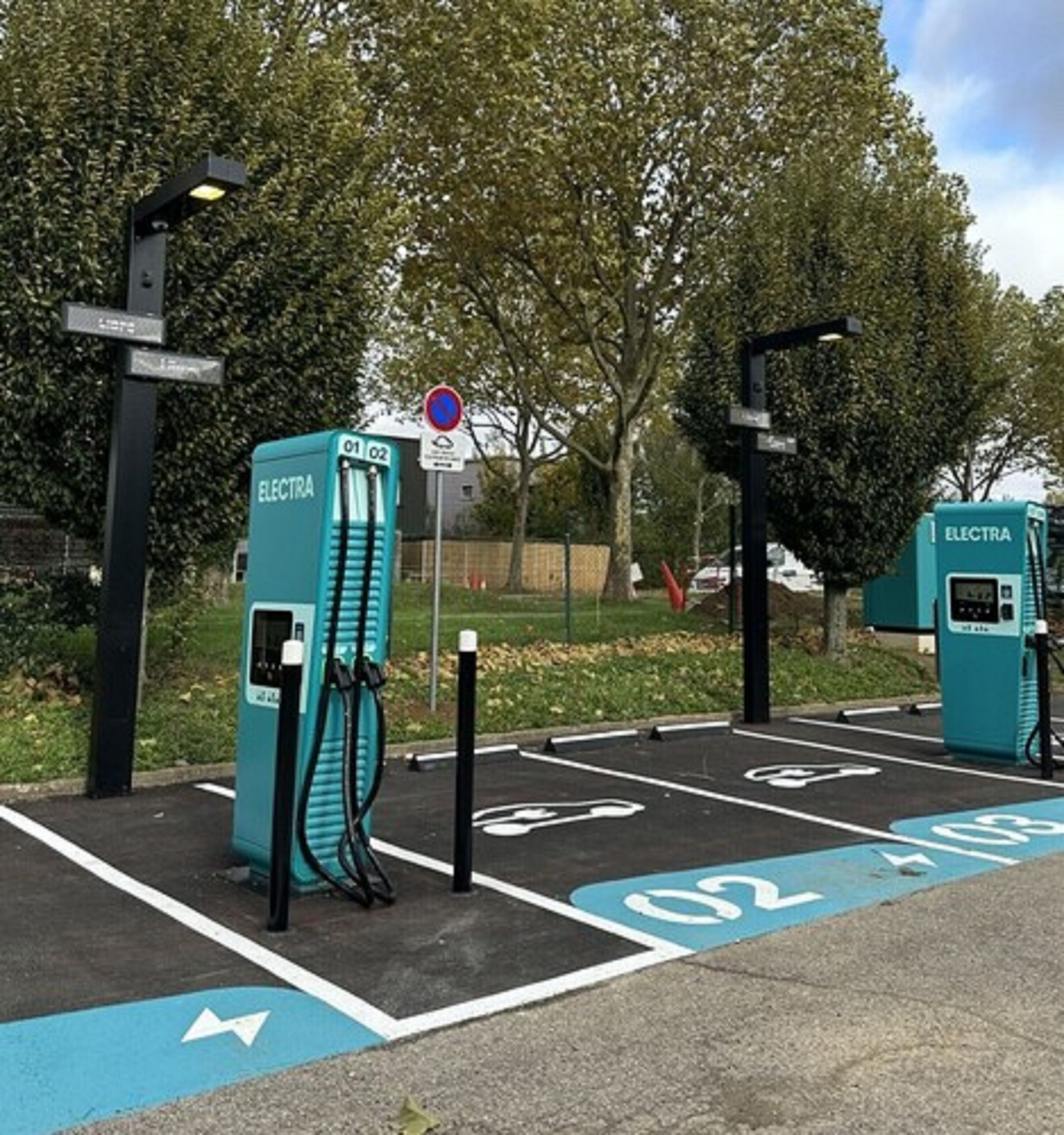 Una estación de carga para vehículos eléctricos con dos puntos de carga, plazas de aparcamiento señalizadas y flechas direccionales en el pavimento.