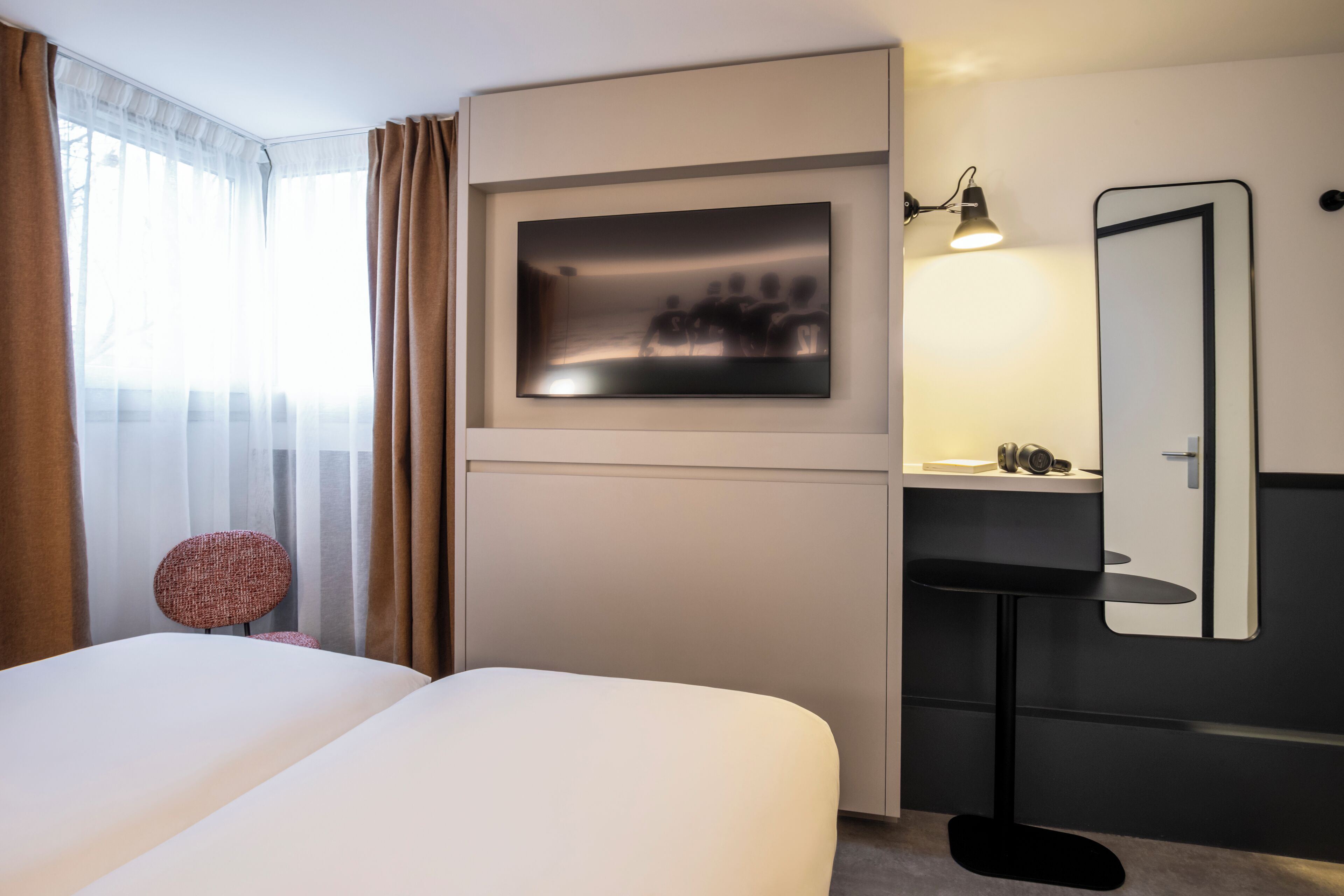 Ein modernes Hotelzimmer mit einem großen Bett, einem an der Wand befestigten Fernseher, einem Nachttisch mit Lampe und einem Spiegel.