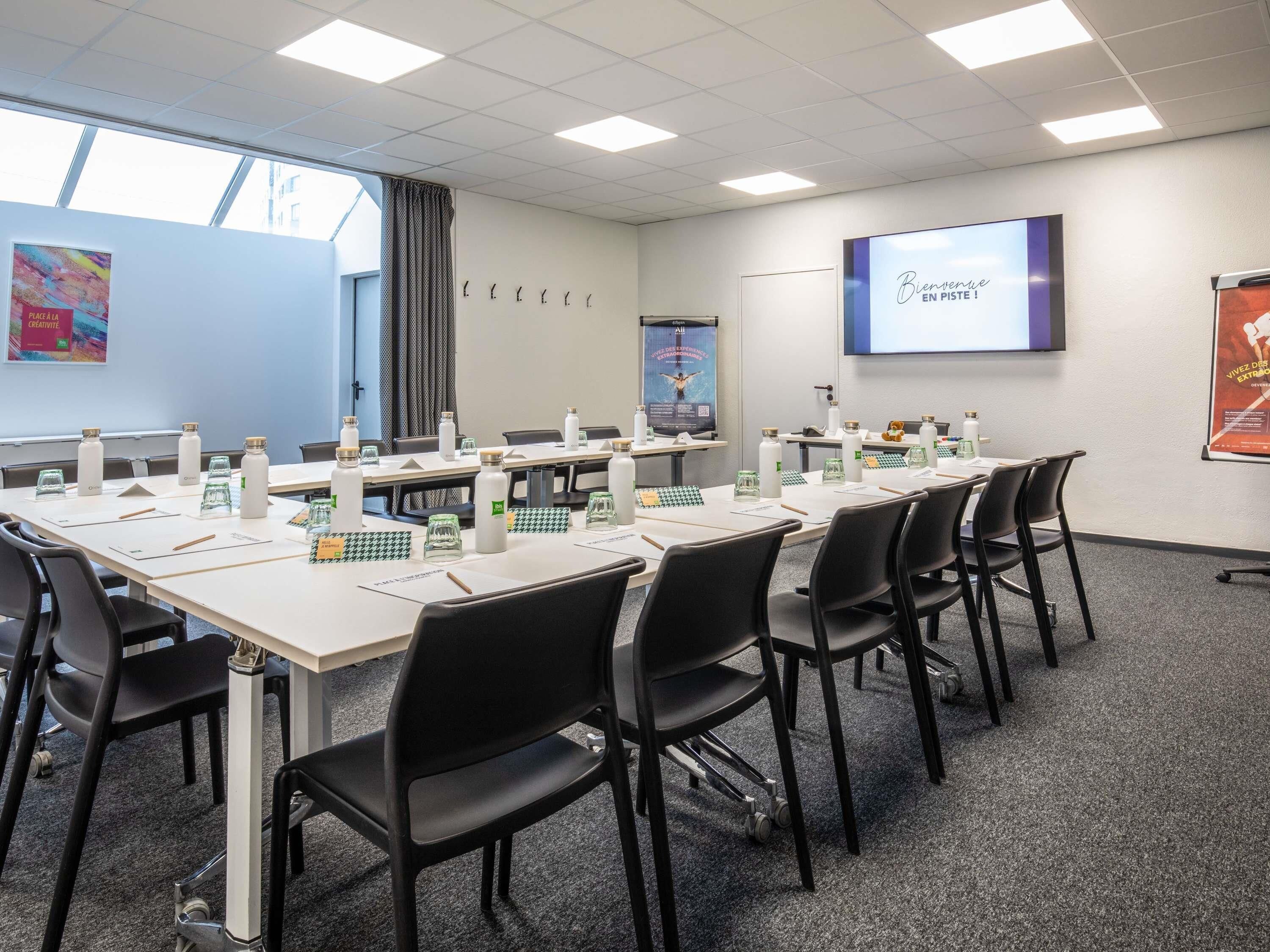 A conference room with tables, chairs, water bottles, and a screen displaying text.