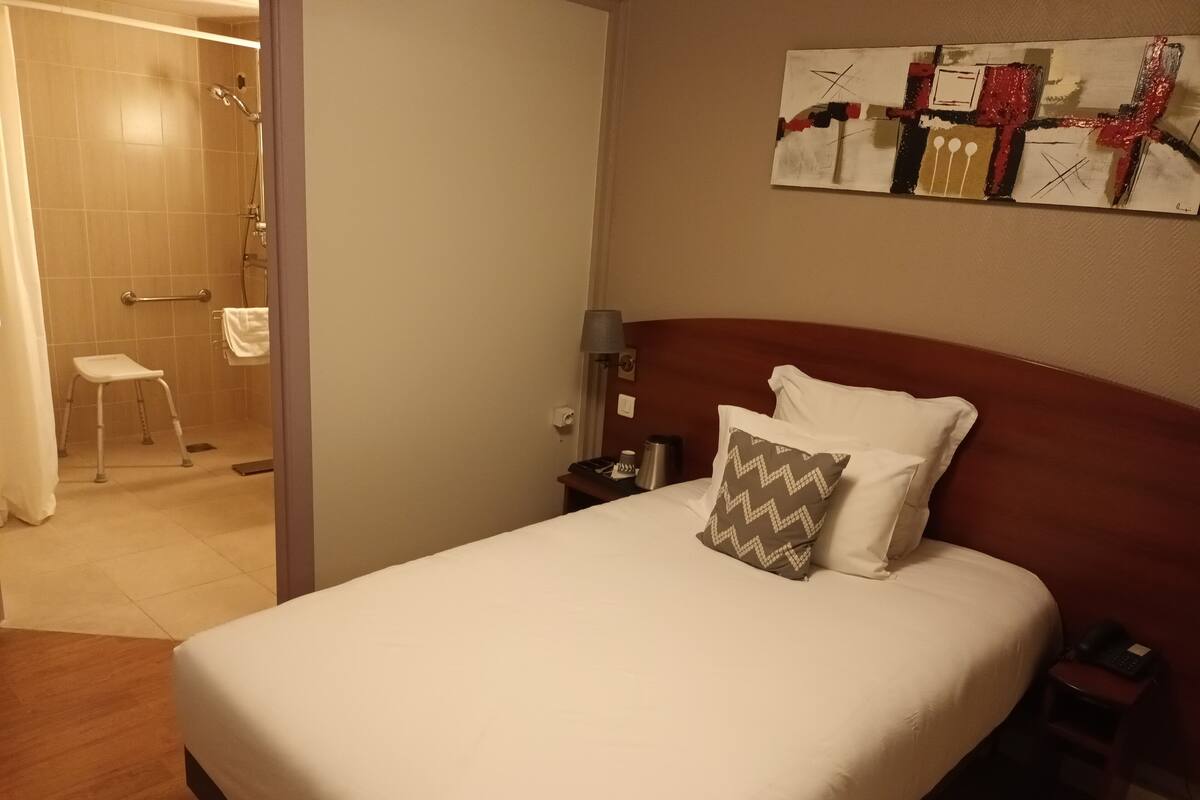 Quarto de hotel com cama, mesa de cabeceira, luminária, chuveiro e banheiro.