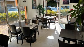 Un espace repas avec des tables et des chaises, de grandes fenêtres et une vue sur les voitures garées et la verdure à l’extérieur.