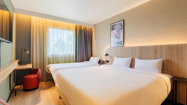 Habitación de hotel con dos camas, un escritorio, una silla roja y un ventanal con cortinas.