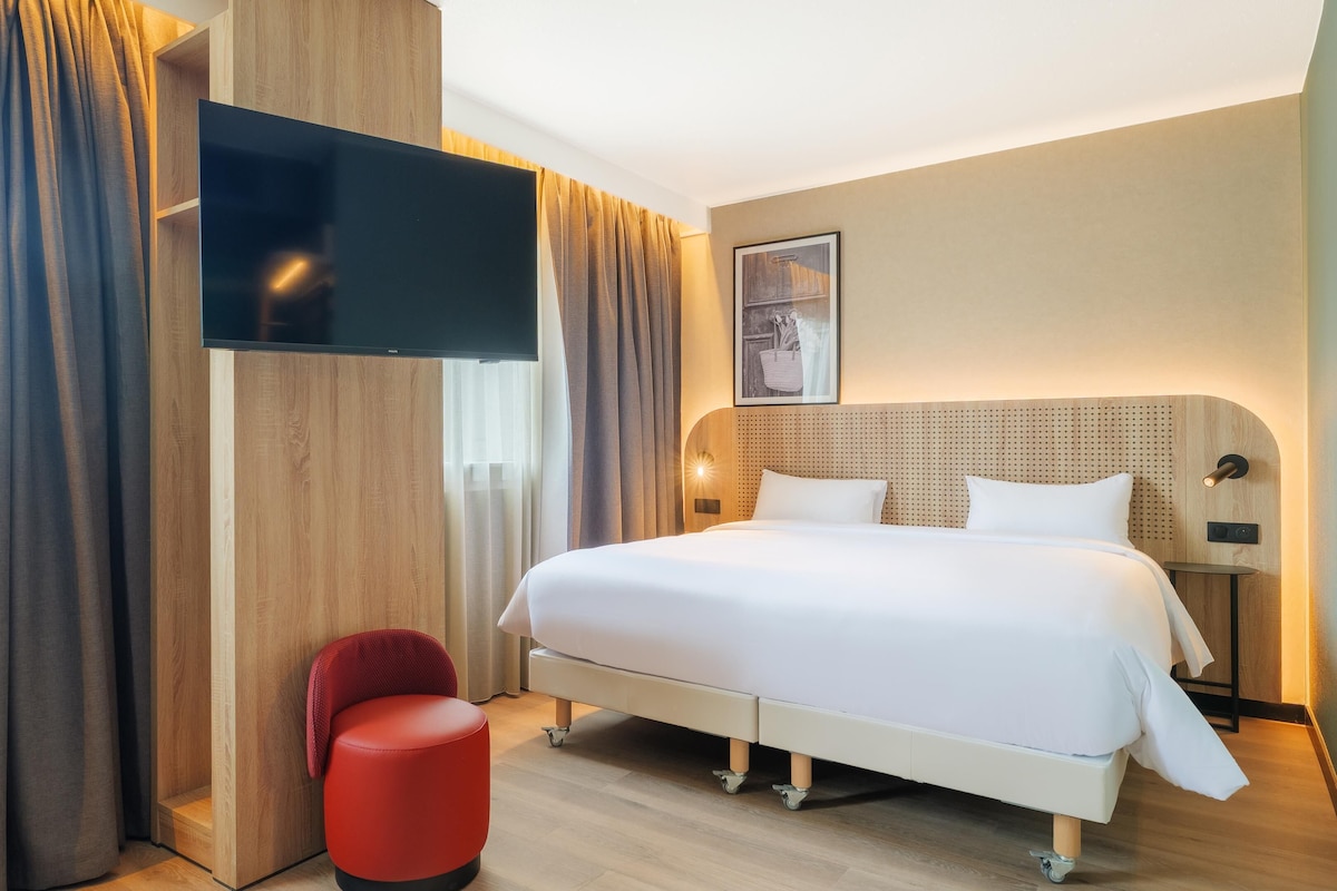 A hotel room with a bed, a TV, a red chair, and a wall-mounted light.