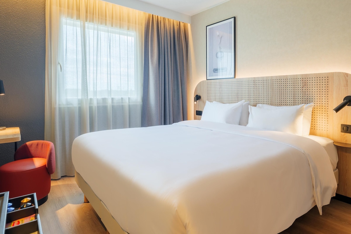 A hotel room with a large bed, a bedside table, a red chair, and a window with curtains.