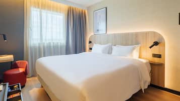 A hotel room with a large bed, a bedside table, a red chair, and a window with curtains.