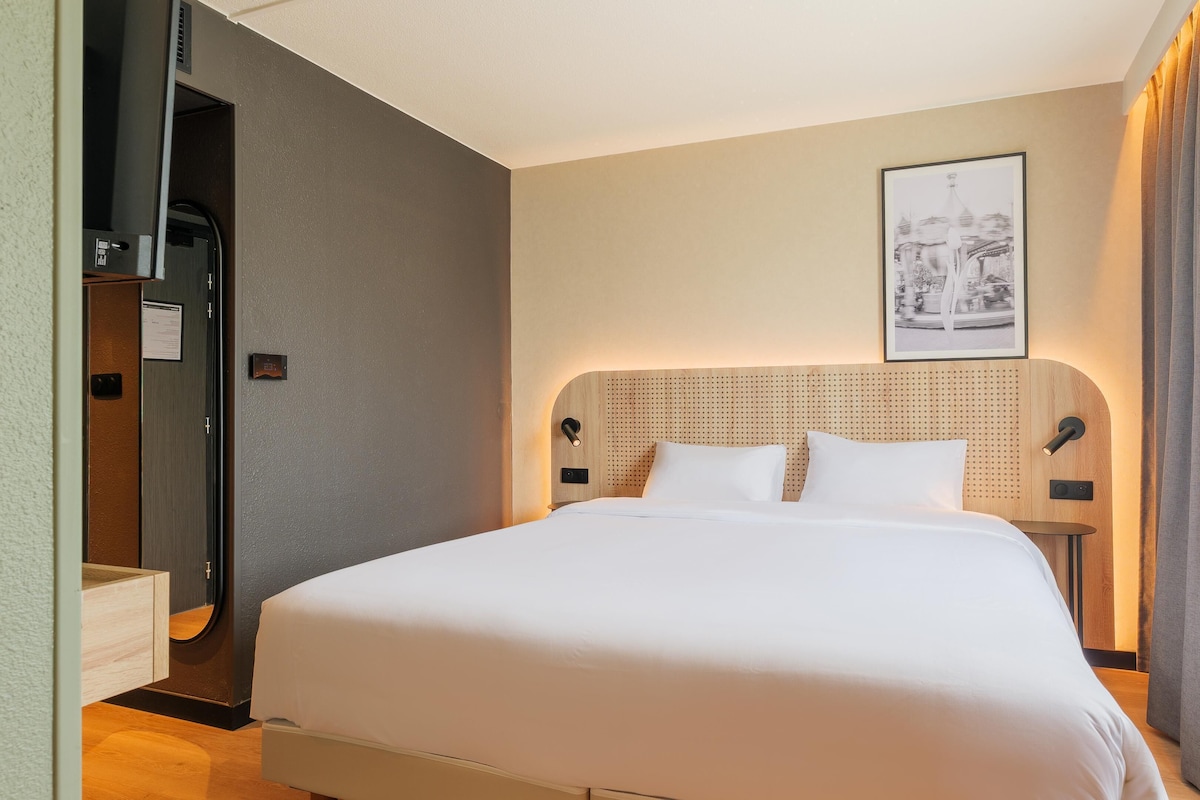 A hotel room with a bed, a headboard with a perforated design, a framed picture on the wall, and a door with a handle.