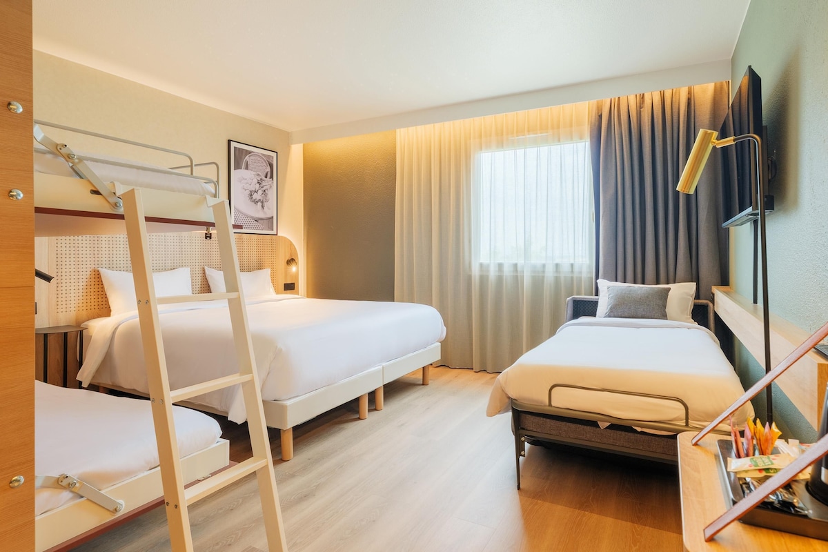 A hotel room with two beds, a bunk bed, a desk, a chair, a TV, and a window with curtains.