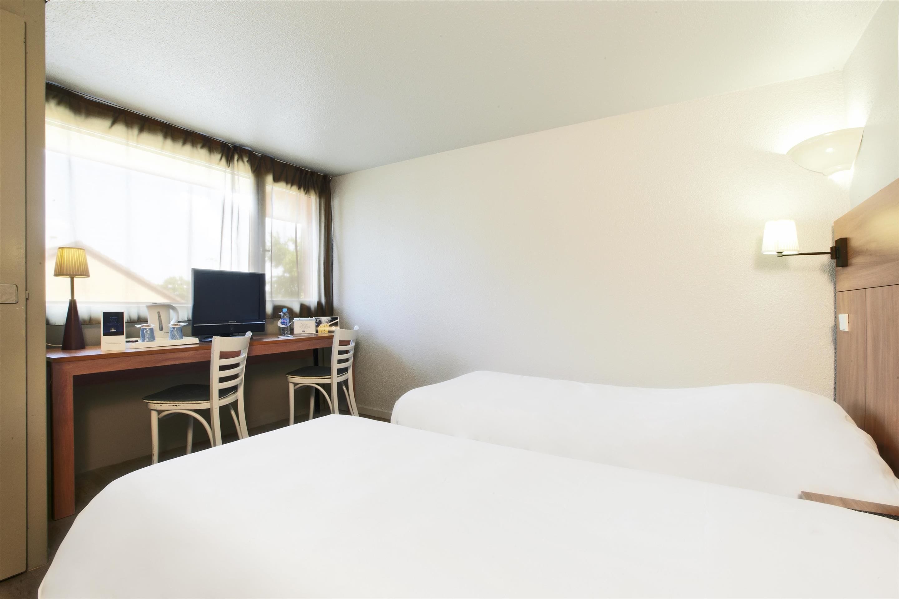 A hotel room with two beds, a desk with a computer, chairs, and a lamp.