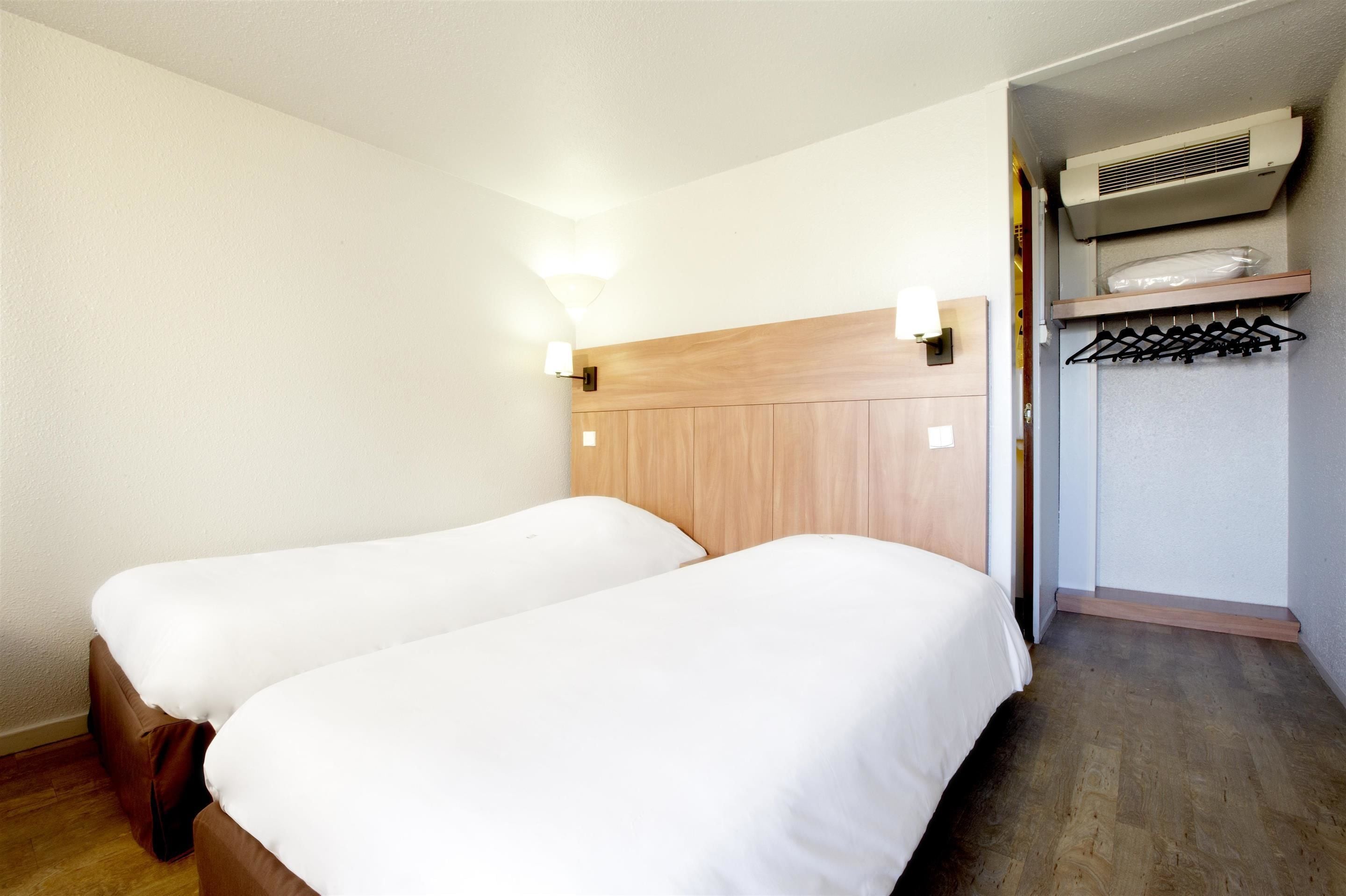 Une chambre d’hôtel avec deux lits simples, une tête de lit en bois, une lampe fixée au mur et un placard intégré avec une tringle pour suspendre les vêtements.