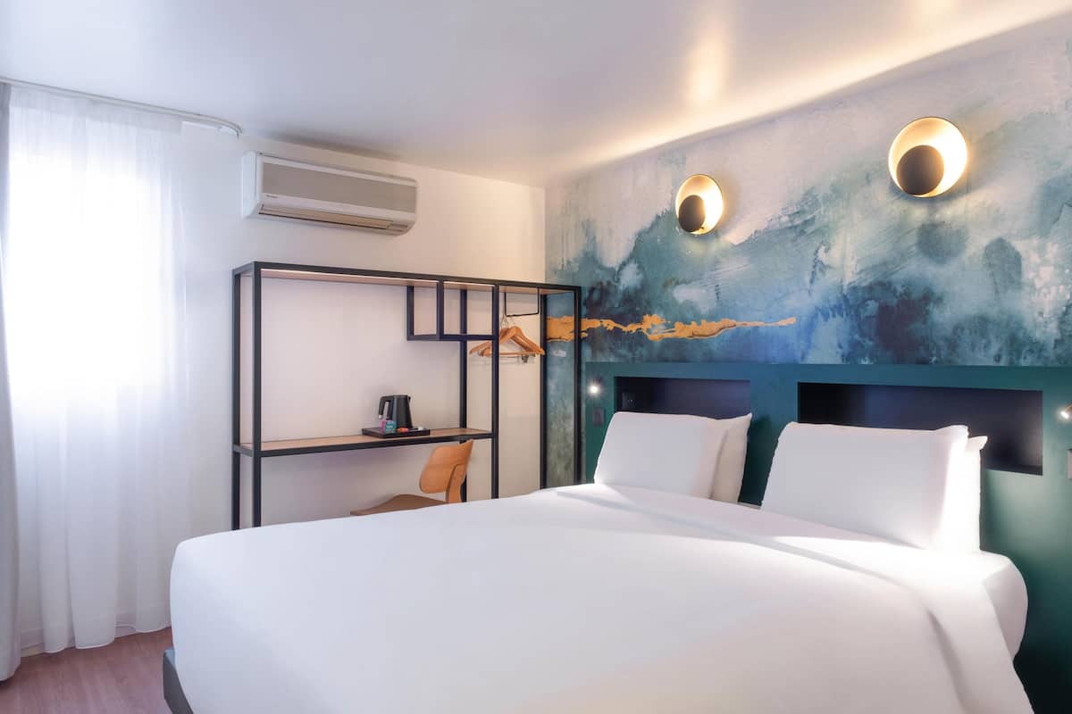 A hotel room with a bed, a desk, and a chair. There is a wall-mounted air conditioner and two wall sconces.