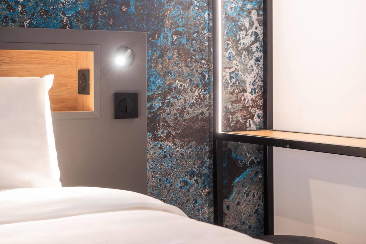 A modern hotel room with a wooden headboard, a white bed, a wall-mounted control panel, and a textured blue and black wall.