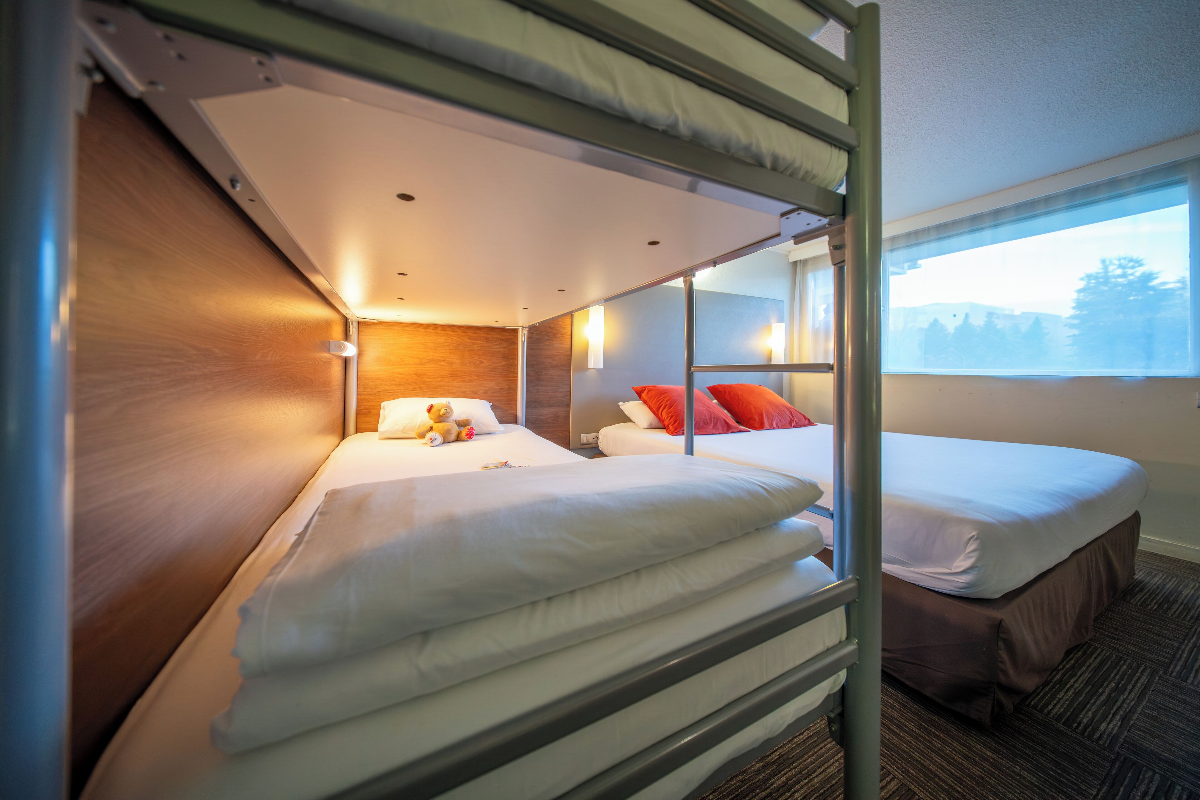 Een slaapkamer met stapelbed, een houten hoofdbord, een raam met uitzicht op bomen, en een bed met witte beddengoed en rode kussens.