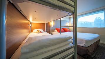Een slaapkamer met stapelbed, een houten hoofdbord, een raam met uitzicht op bomen, en een bed met witte beddengoed en rode kussens.