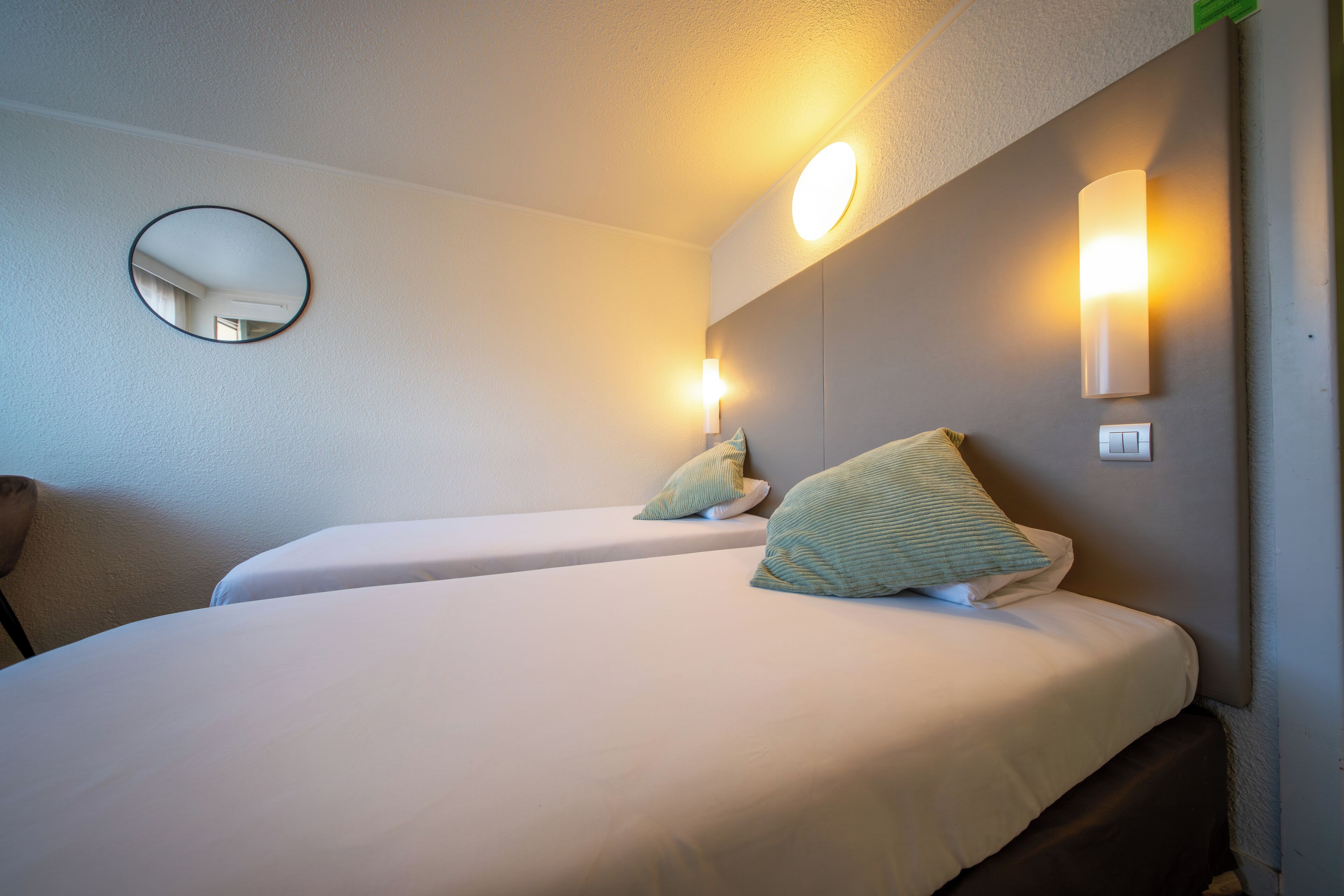 Una camera d'albergo con due letti, uno specchio rotondo e luci a parete.
