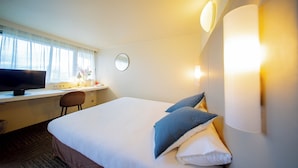 Een hotelkamer met een bed, een bureau met een computer, een stoel, een ronde spiegel en wandlampen.