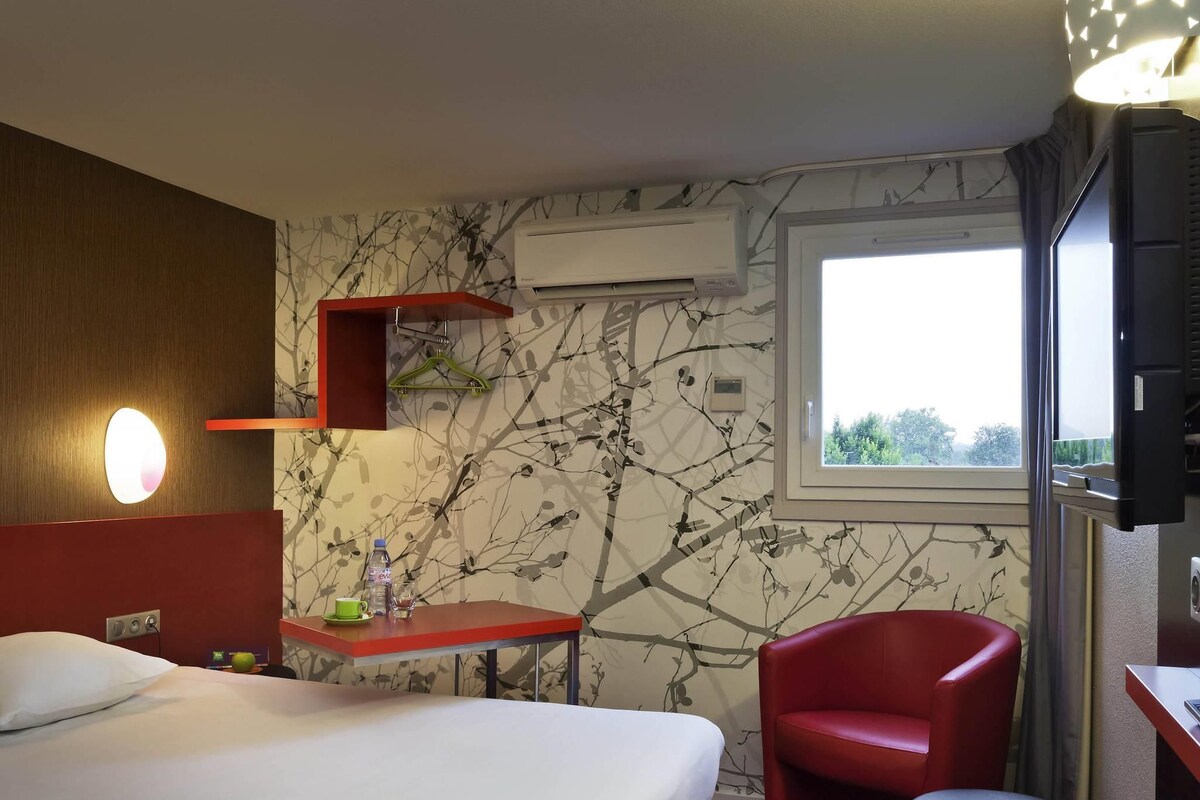 Ein Hotelzimmer mit einem Bett, einem roten Sessel, einem kleinen Tisch mit Flasche und Glas, einer Wand-Klimaanlage und einem Fenster mit Blick.