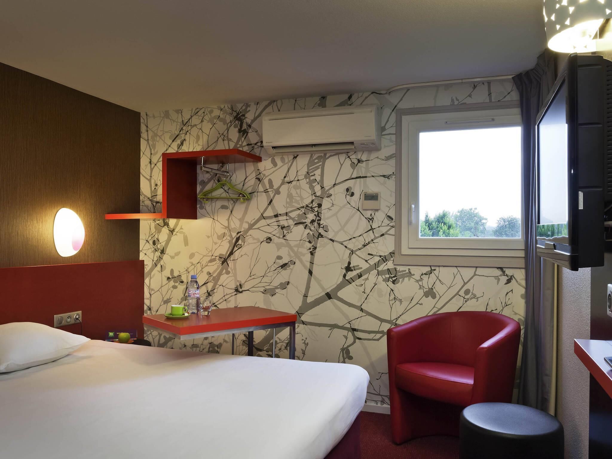A hotel room with a bed, a red chair, a small table with a bottle and glass, a wall-mounted air conditioner, and a window with a view.