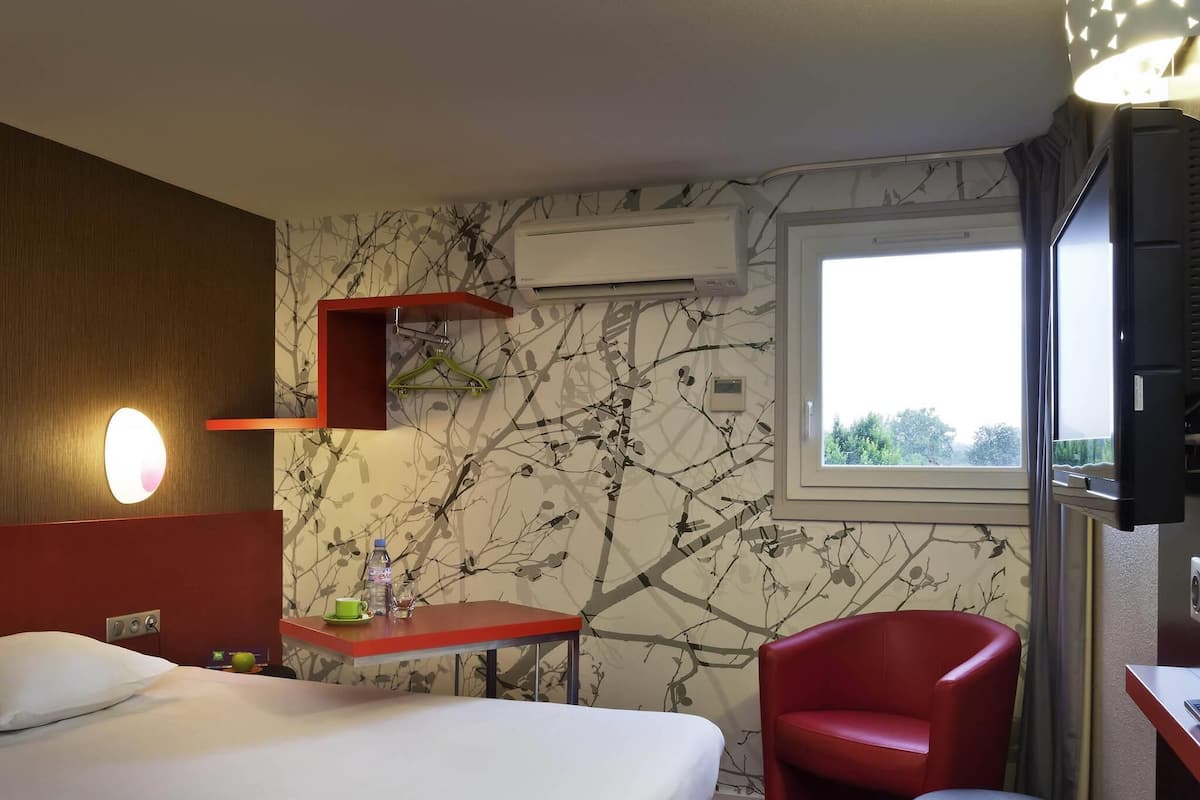 Habitación de hotel con cama, una silla roja, una mesita con botella y vaso, aire acondicionado de pared y ventana con vistas.