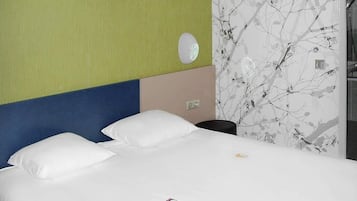 A hotel room with a bed, white pillows, a wall clock, and a wall with a branch pattern.