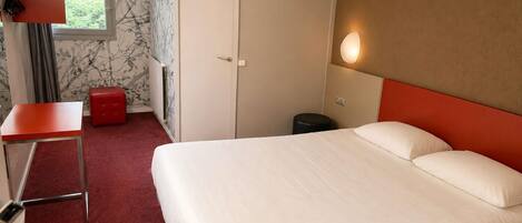 Habitació d'hotel amb llit, escriptori, cadira, tamboret vermell, radiador, finestra i llum penjada a la paret.