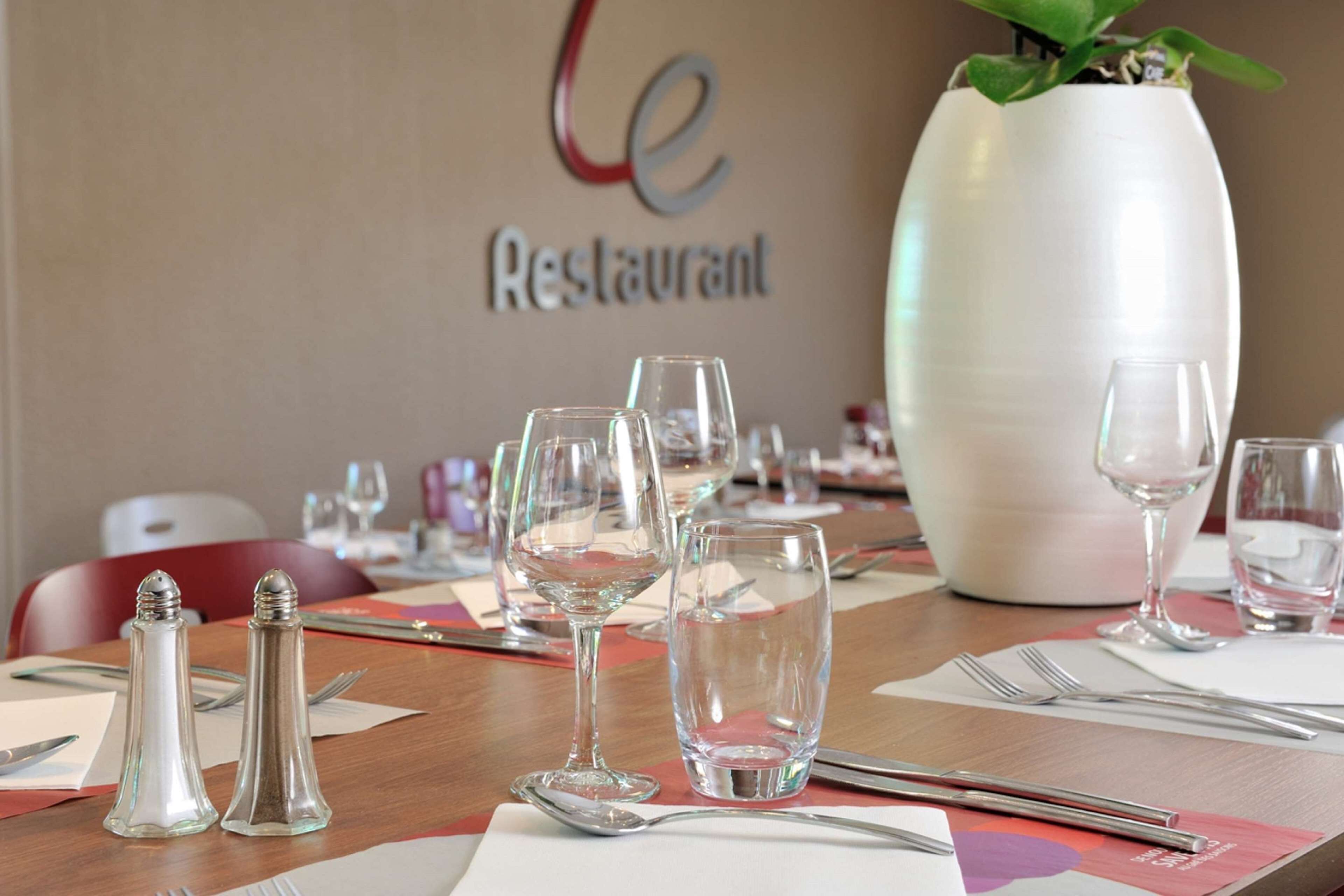 Una mesa de comedor dispuesta con copas de vino, cubiertos y saleros y pimenteros, con una planta en maceta y un letrero que dice "Le Restaurant" al fondo.