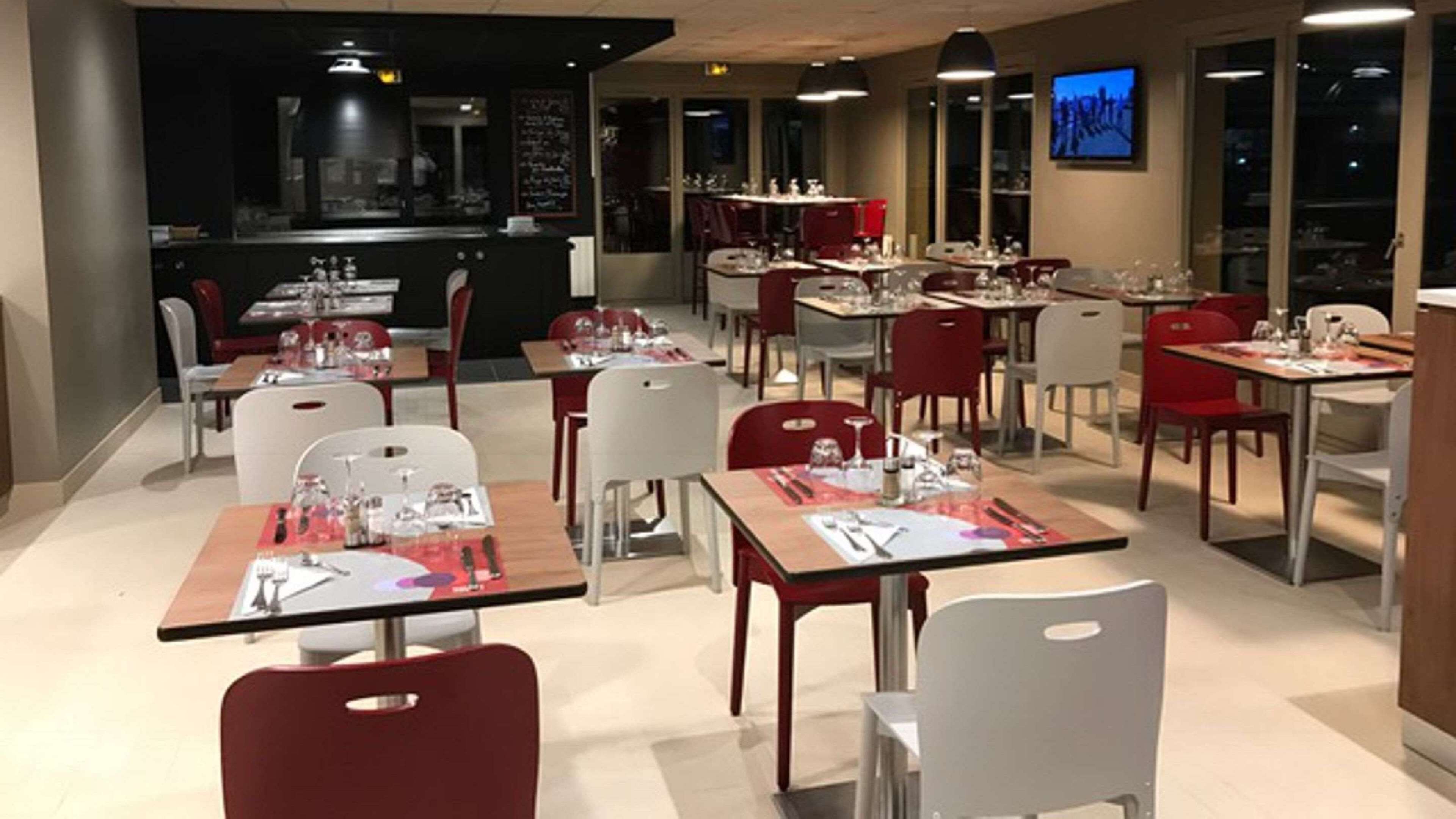 En restaurant med borde dækket til spisning, røde og hvide stole, og et barområde i baggrunden.