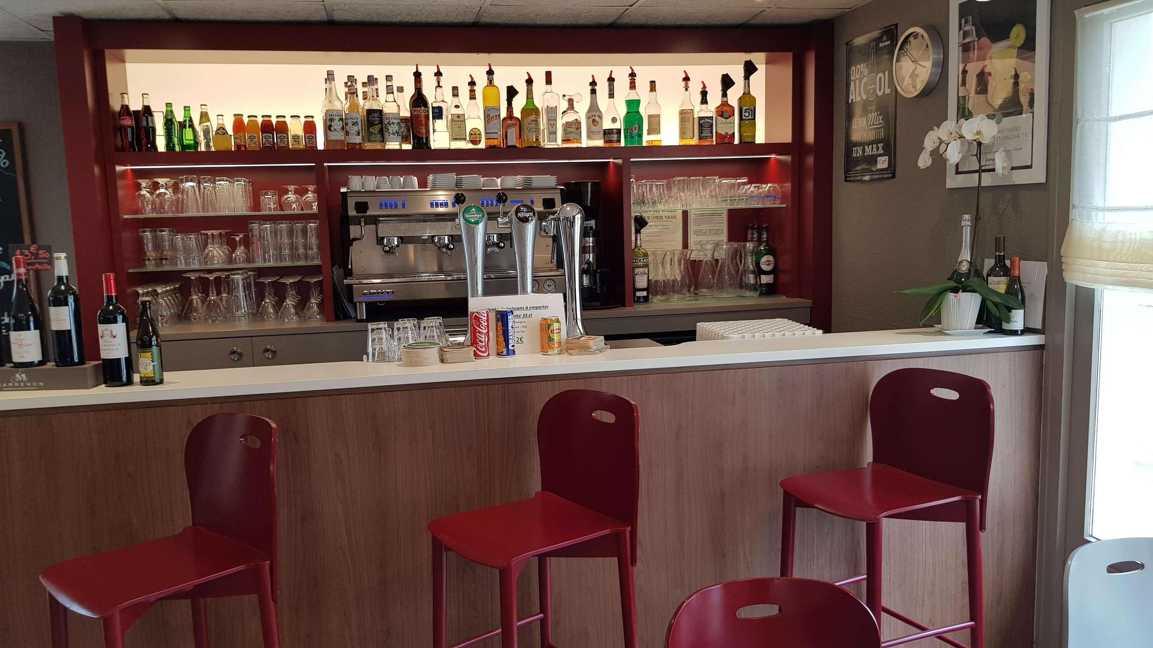 Een bar met een toonbank, flessenkasten vol drank, glazen en een koffieapparaat. Voor de toonbank staan drie rode barkrukken.