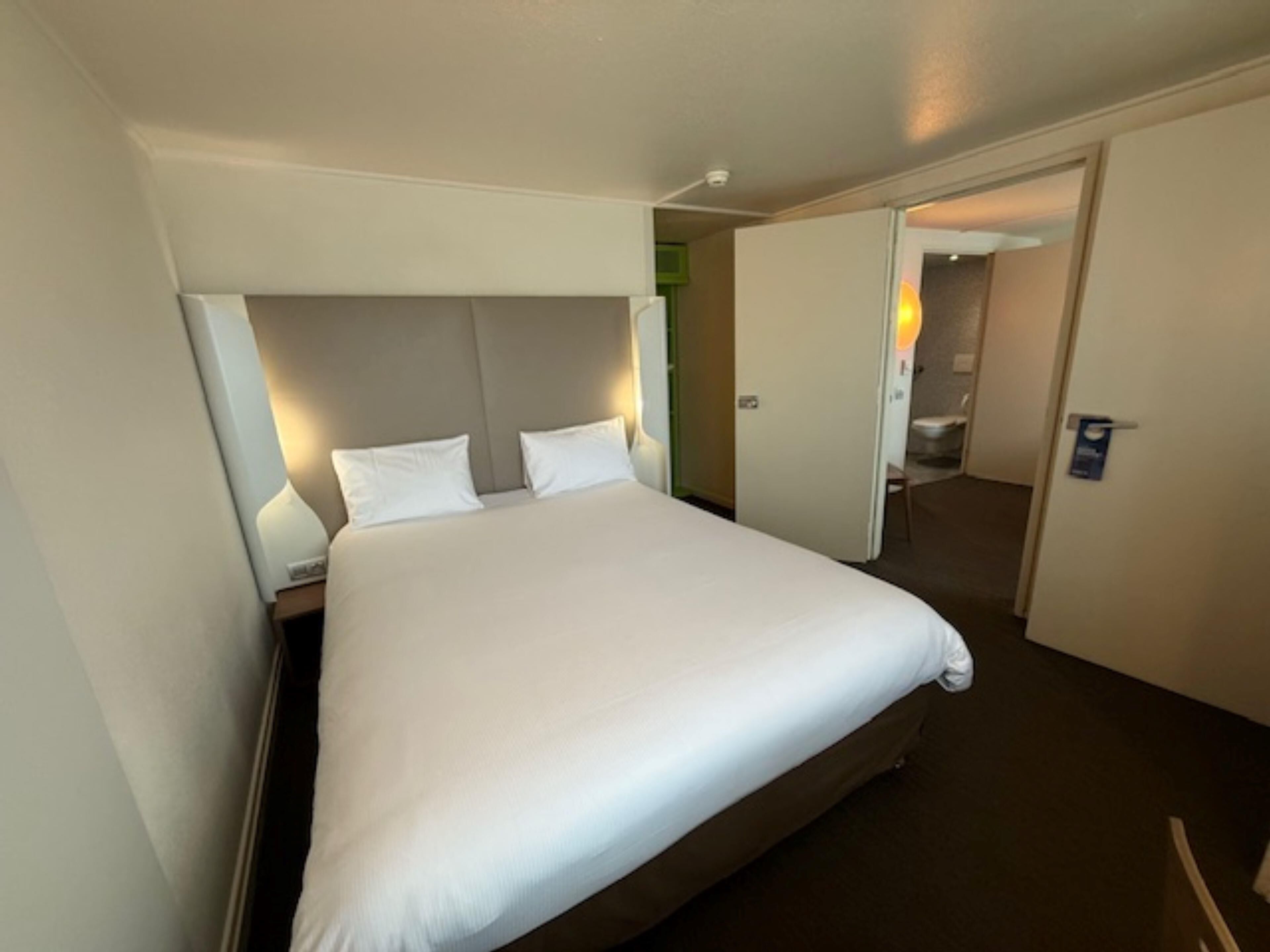 A hotel room with a large bed, a nightstand, a wall-mounted lamp, and a door leading to a bathroom.