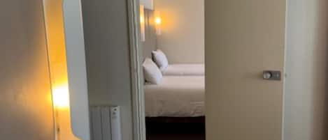 Una habitación de hotel con una cama, una lámpara de noche, un radiador y una puerta que da a otra habitación.