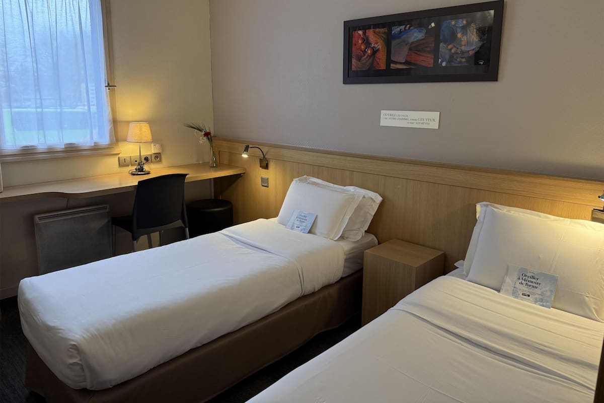 Quarto de hotel com duas camas de solteiro, uma escrivaninha com cadeira, uma mesa pequena e um quadro na parede.