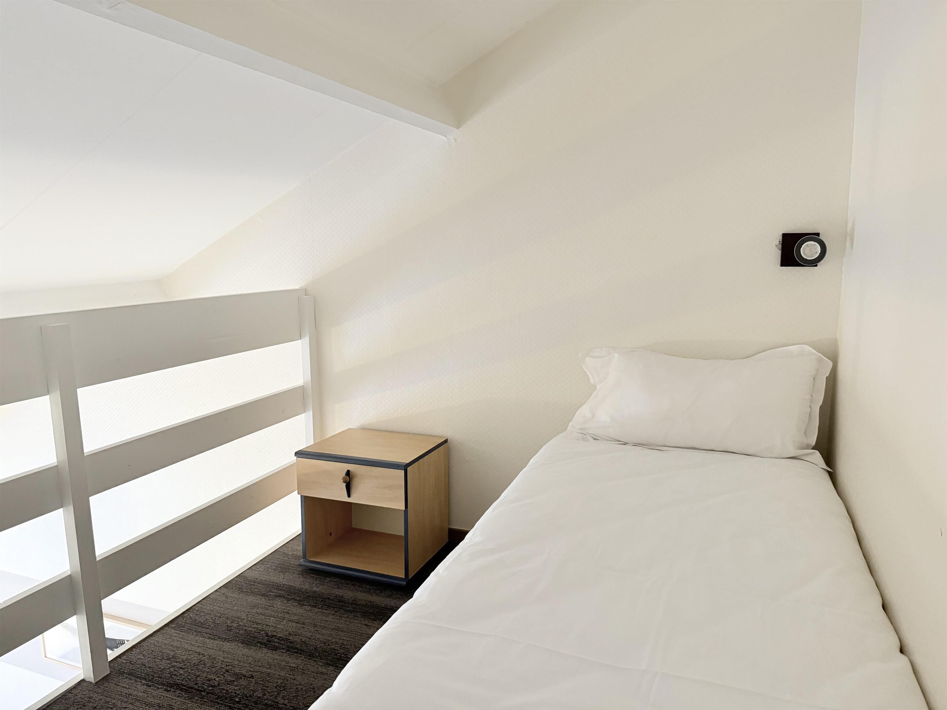 Een kleine, eenvoudige kamer met een bed, een nachtkastje en een raam.
