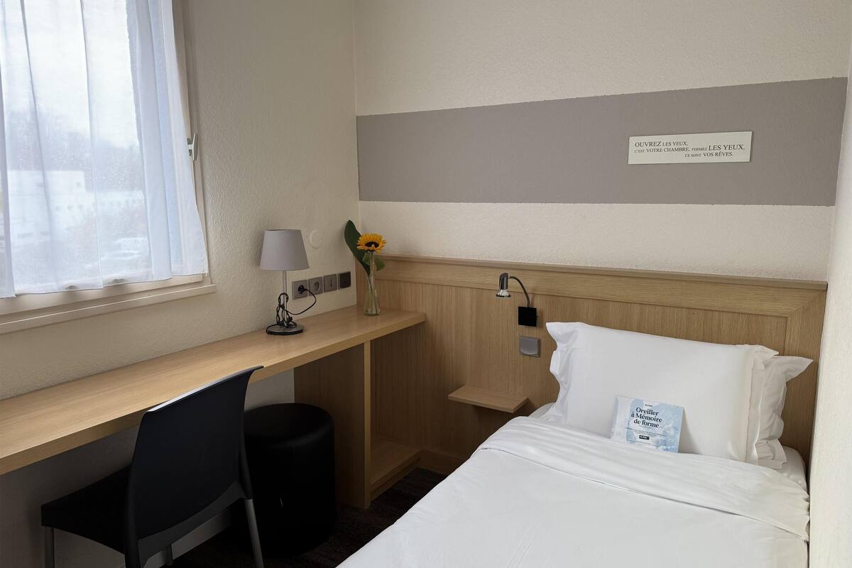 Quarto de hotel com uma cama, escrivaninha, cadeira, abajur, vaso de flores e uma placa na parede.